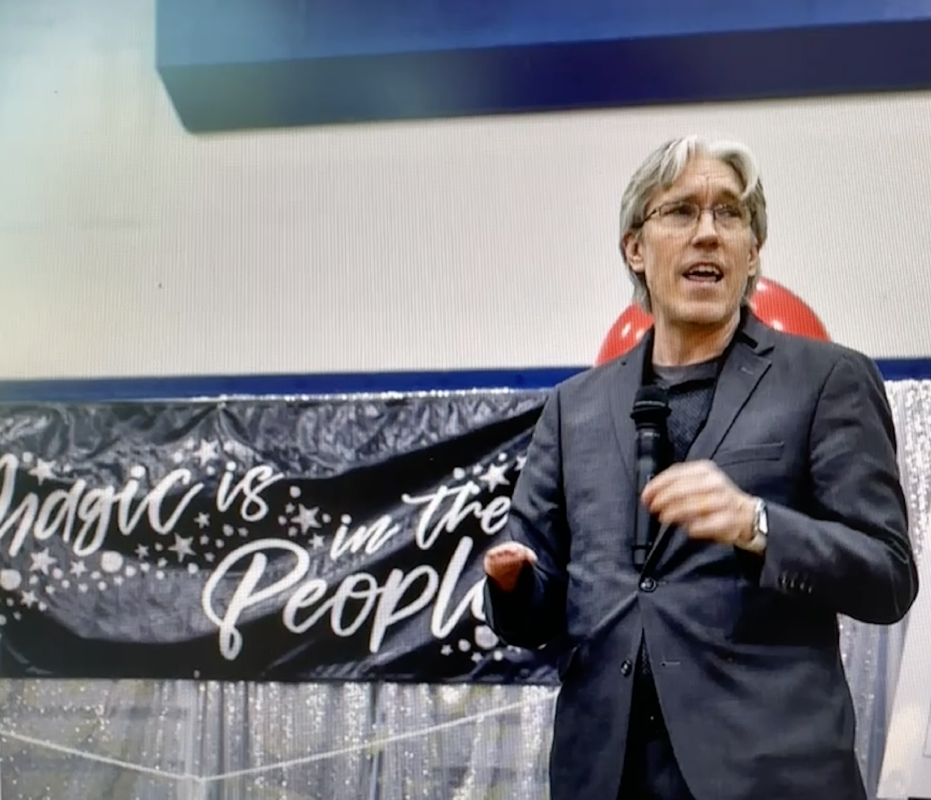 Chris Canfield speaking in front of a “magic is in the people” banner